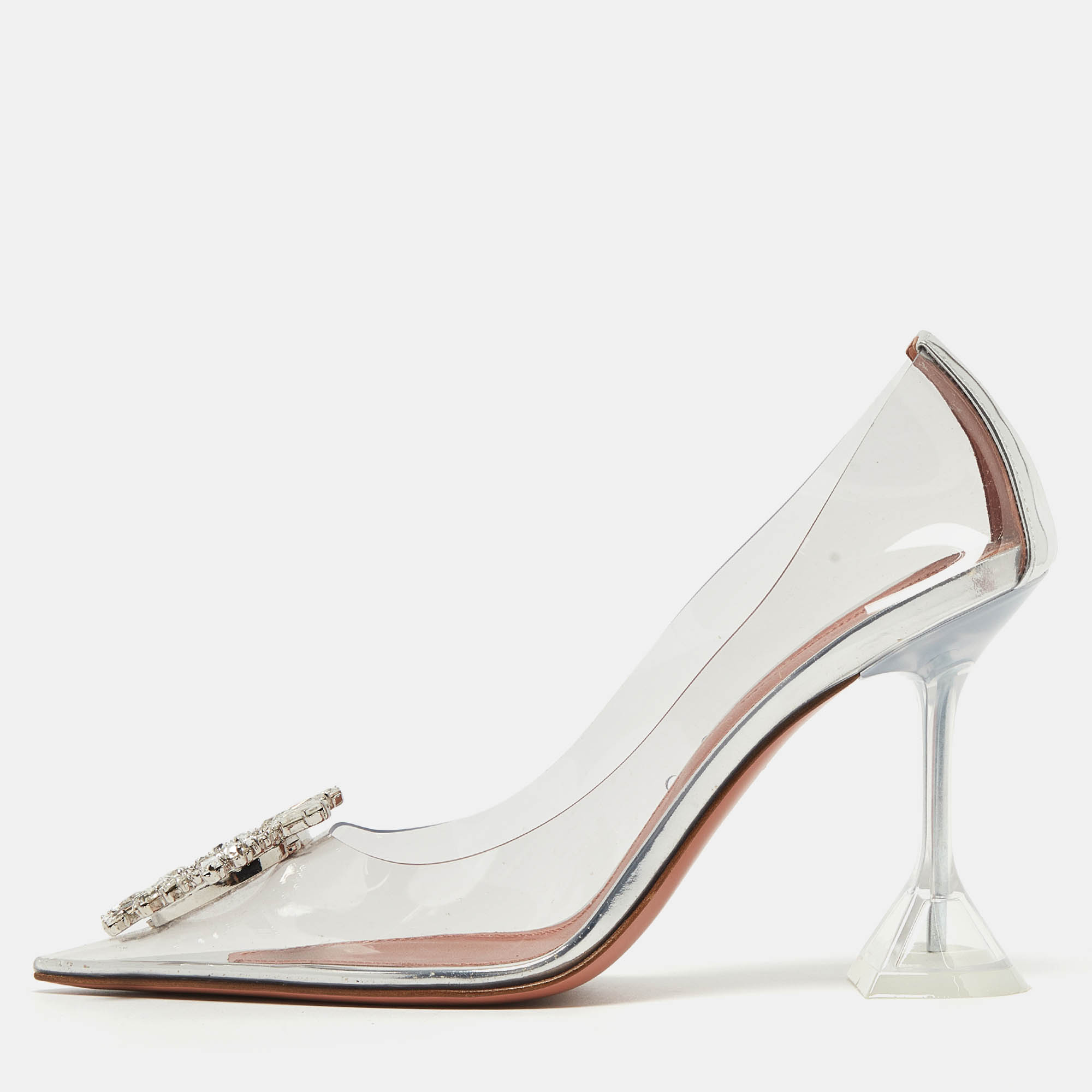The Amina Muaddi Begum pumps are striking high heeled shoes. They feature transparent PVC uppers a pointed toe and a sculpted heel with a signature flared design. These pumps exude modern elegance combining transparency with a bold embellishment for a unique and stylish look.