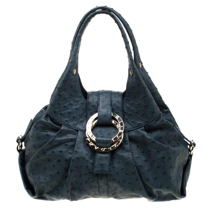 A known style from the house of Bvlgari Chandra is loved for its comfortable silhouette and elegant appeal. This blue one is meticulously crafted from the exotic ostrich skin and has the signature slouchy shape with broad pleats dual top handles and a front slender strap which carries twin logo engraved rings in gold tone hardware. The bag boasts of a spacious canvas lined interior which has enough space for all your day to night basics along with a zip pocket for little essentials. Complete with folded corners accented by buckles this hobo is certainly a premium choice for everyday use.