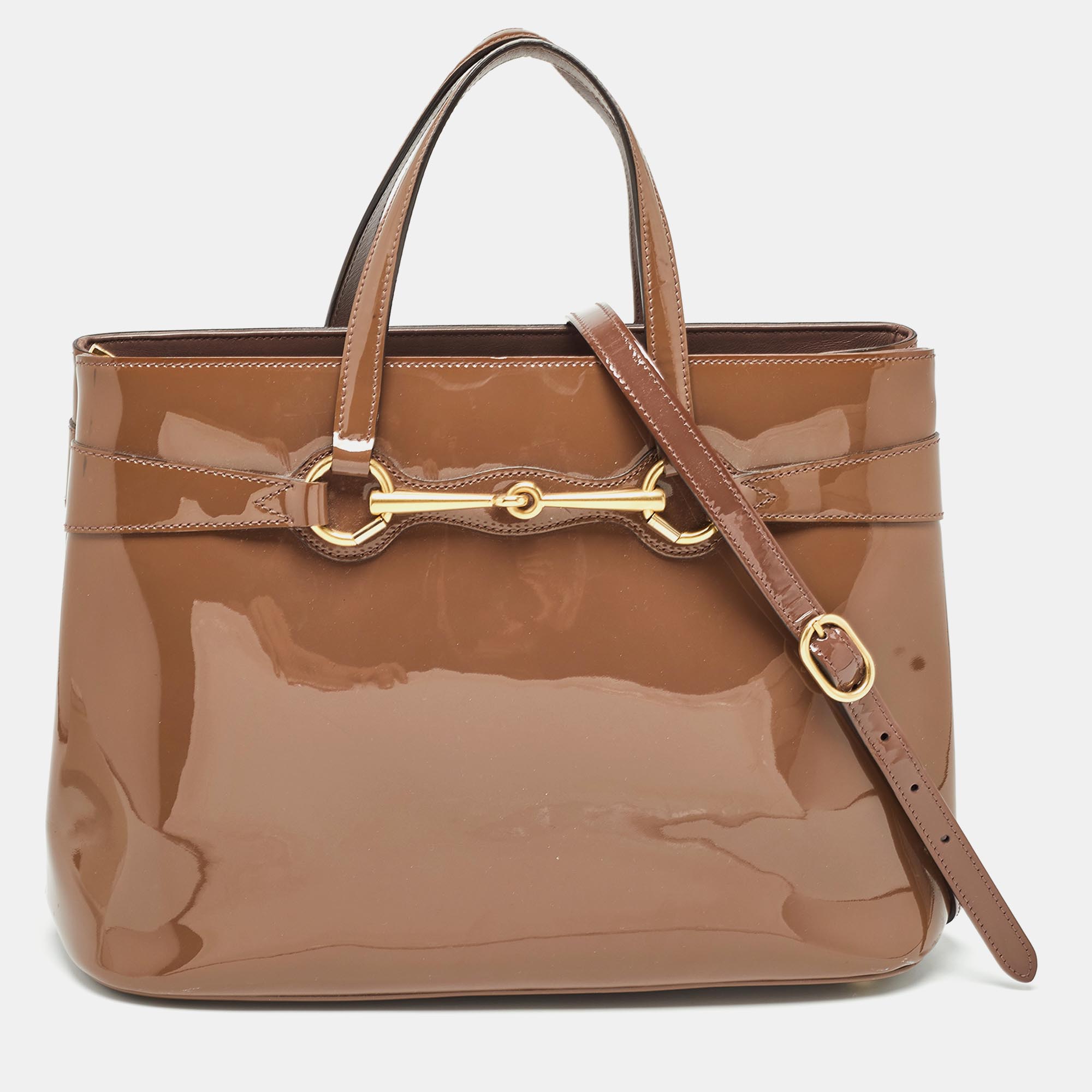 Complete an elegant look with this beautiful Gucci Horsebit tote. It is crafted from patent leather and has the iconic Horsebit accent in gold tone on the front. The brown bag opens to a canvas lined interior that will easily hold all your essentials. This bag can be paraded using the double top handles as well as the optional shoulder strap.